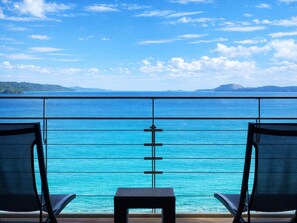 A balcony with two chairs and a small table, overlooking a turquoise sea and distant land under a clear sky.
