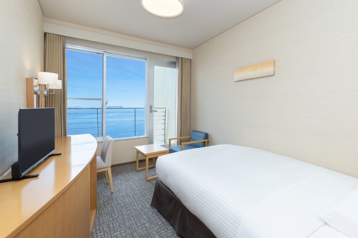 A hotel room with a bed, a desk, a chair, a TV, and a window with a view of the sea.