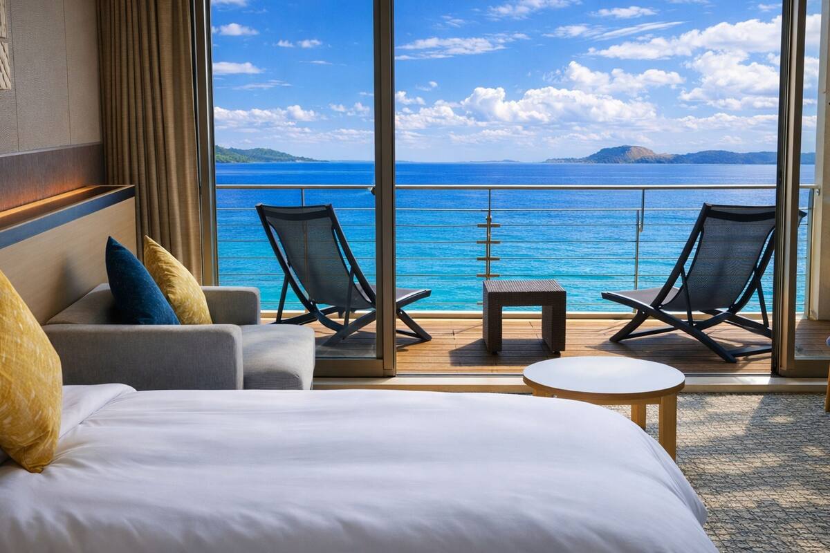 Ein Hotelzimmer mit einem großen Bett, zwei Sessel, einem kleinen Tisch und Blick auf das Meer und das ferne Land.