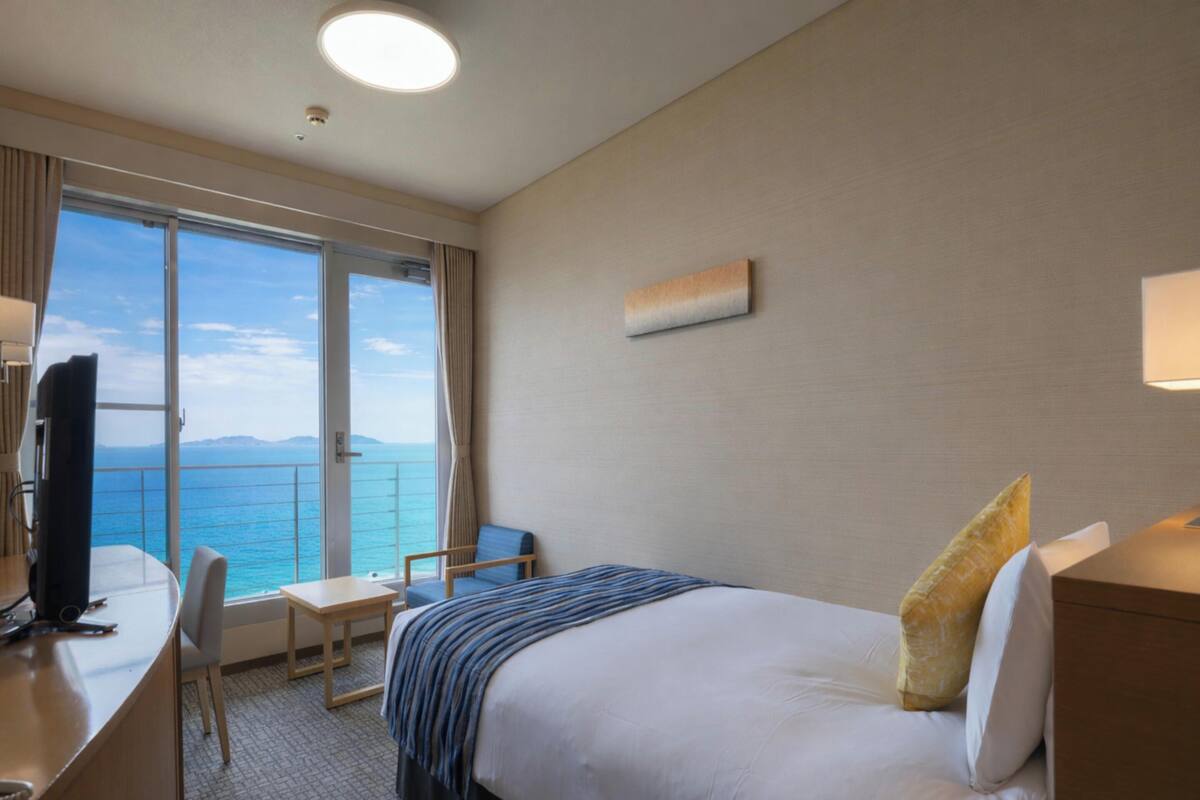 Ein Hotelzimmer mit Bett, Schreibtisch, Stuhl und Fernseher. Ein großes Fenster bietet Blick auf das Meer und die Berge.