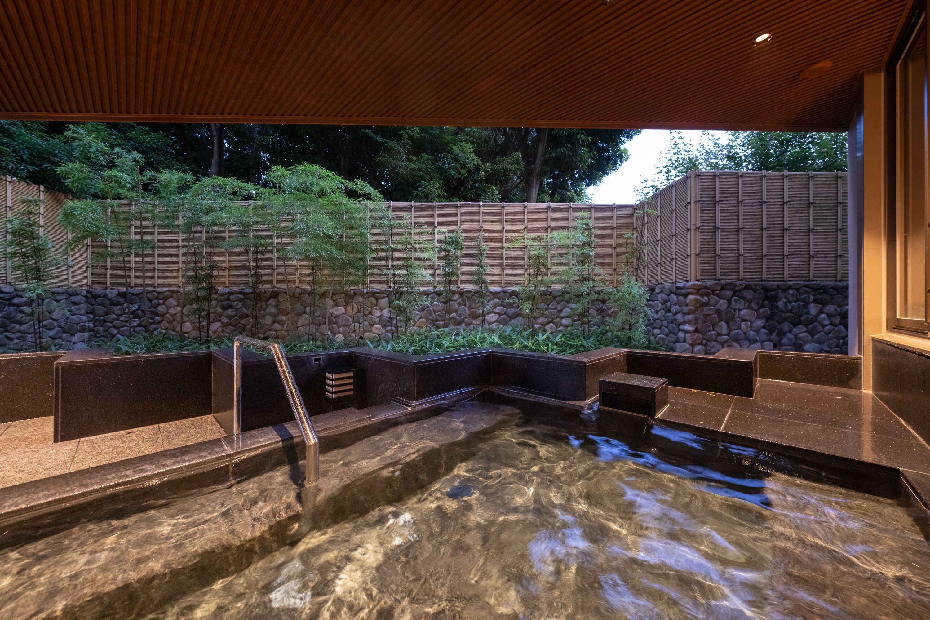 A hot spring area with stone steps leading into a clear pool, surrounded by bamboo plants and a wooden fence.