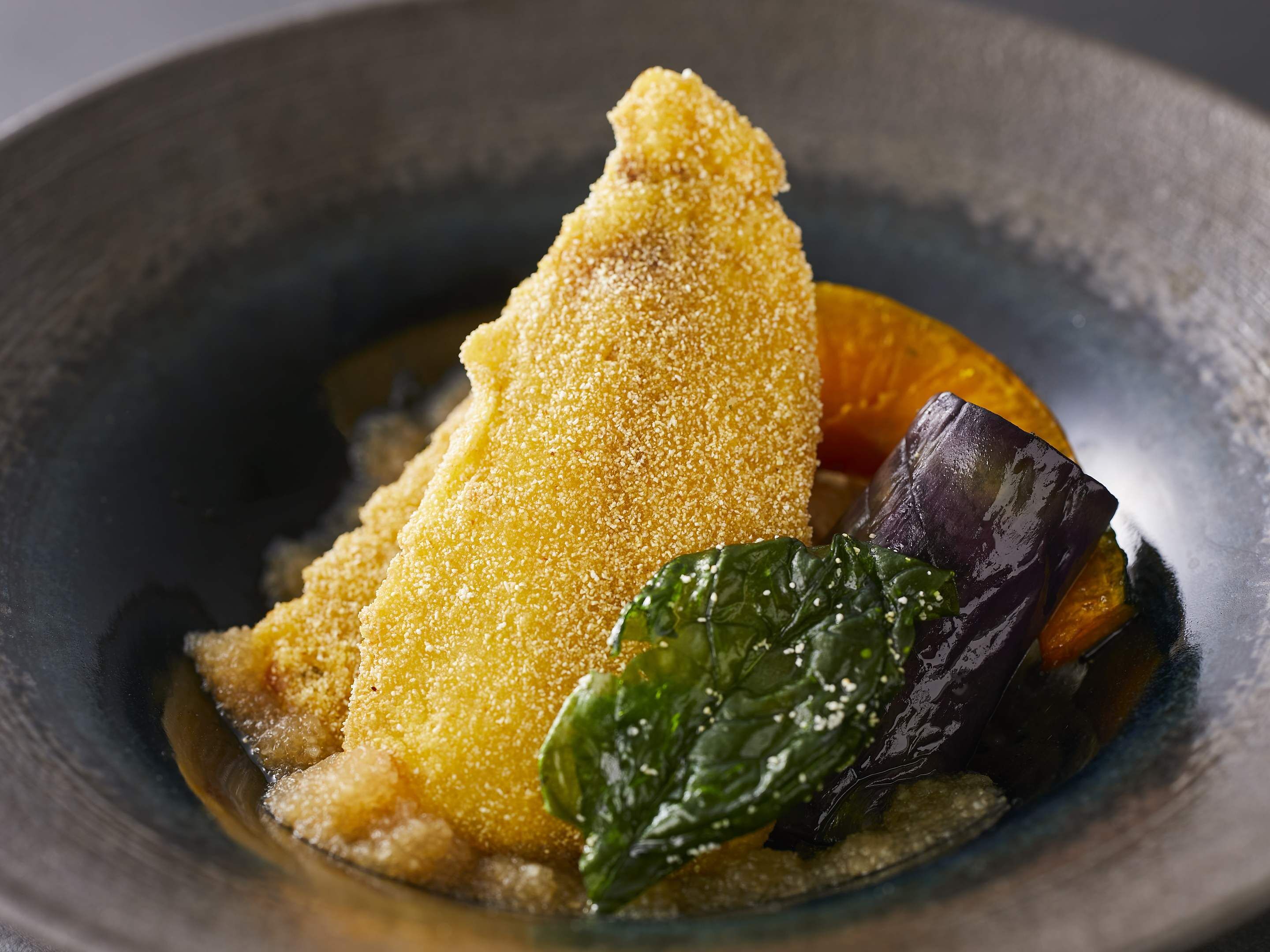 A dish with breaded food, possibly chicken, accompanied by greens and a slice of orange.