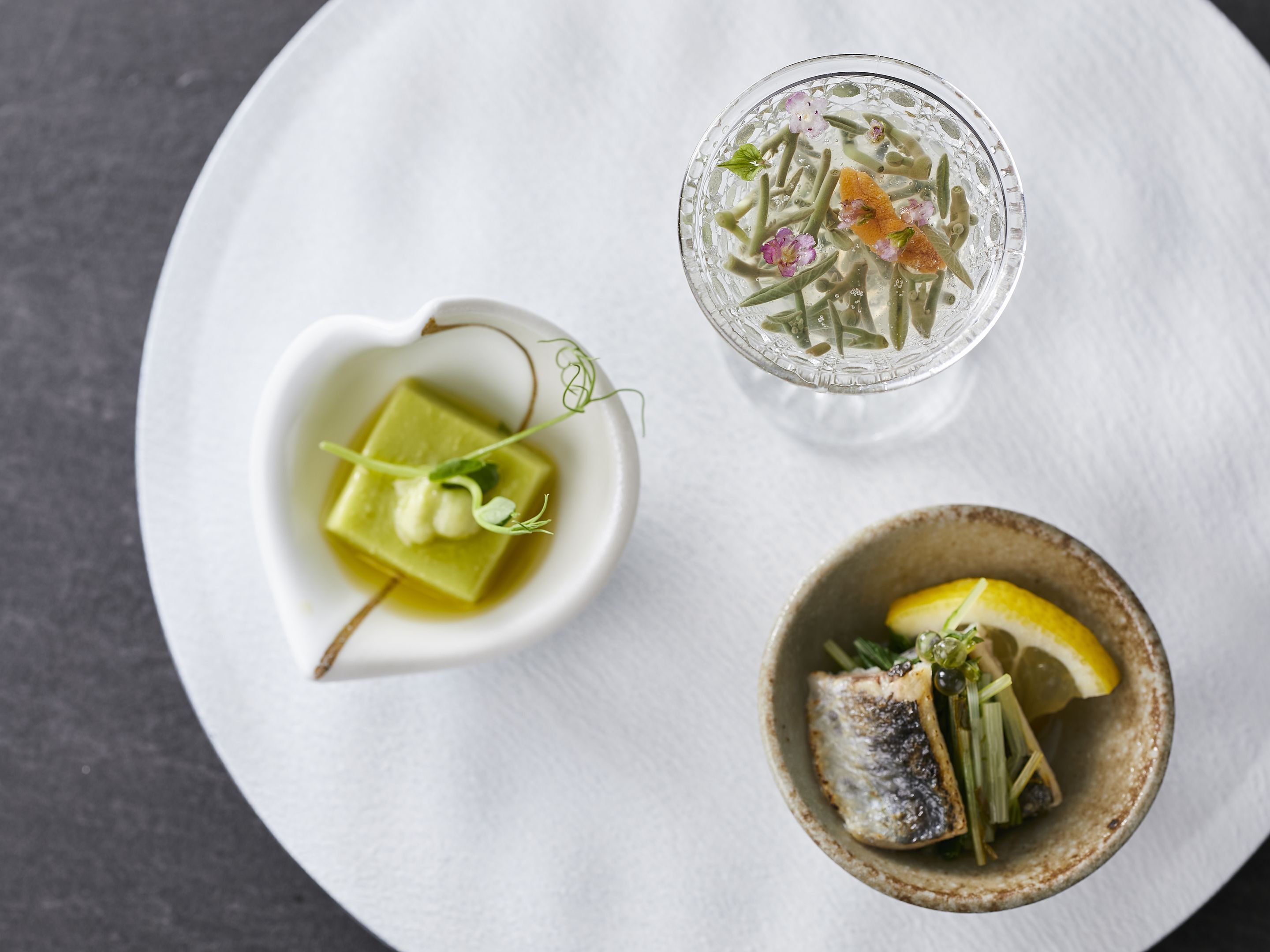 A white tray with three dishes: a heart-shaped bowl with a green square, a clear glass with green beans and flowers, and a small bowl with fish and lemon.