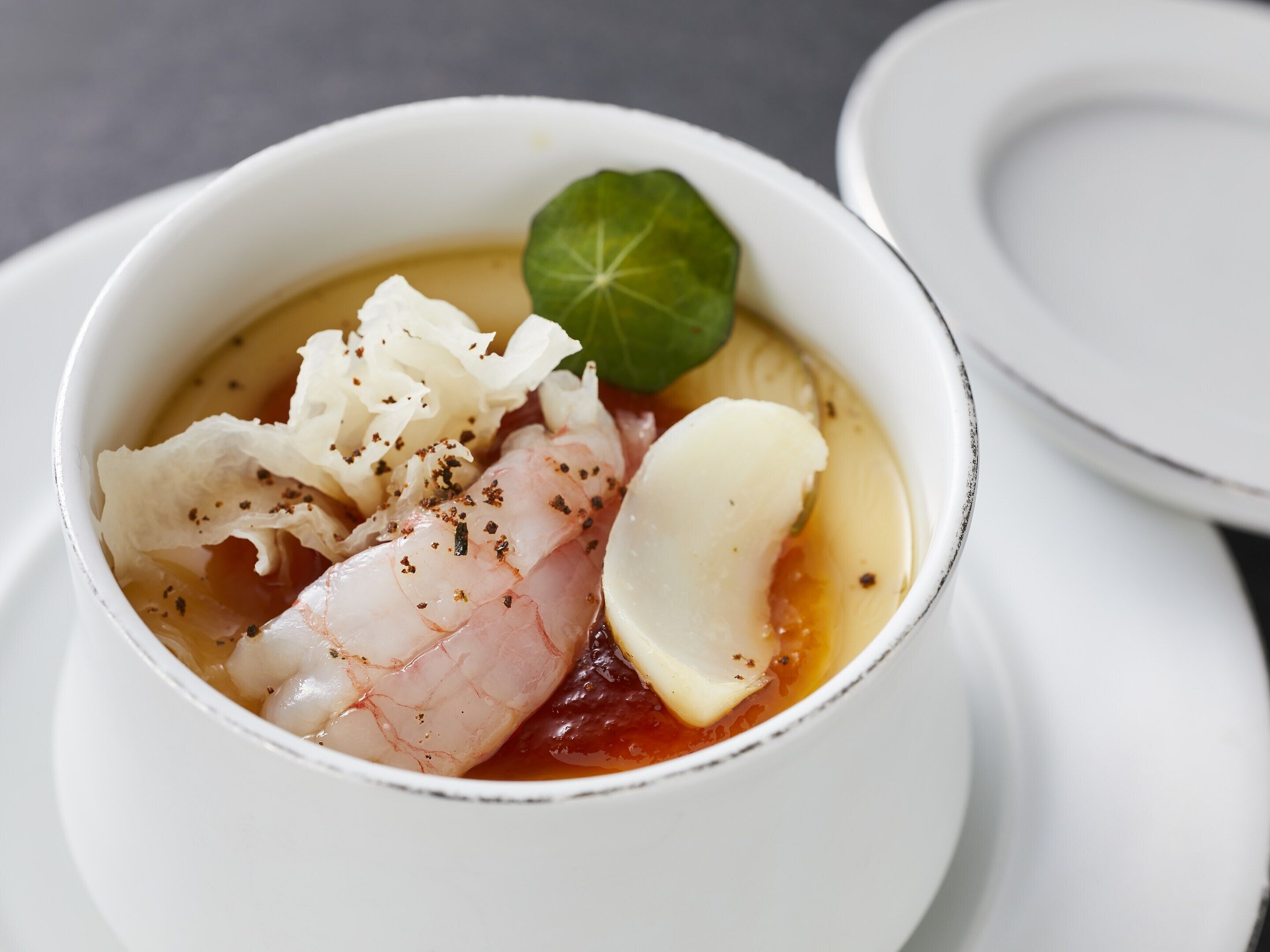Un bol de soupe aux fruits de mer, garni d’une feuille, posé sur une assiette blanche.
