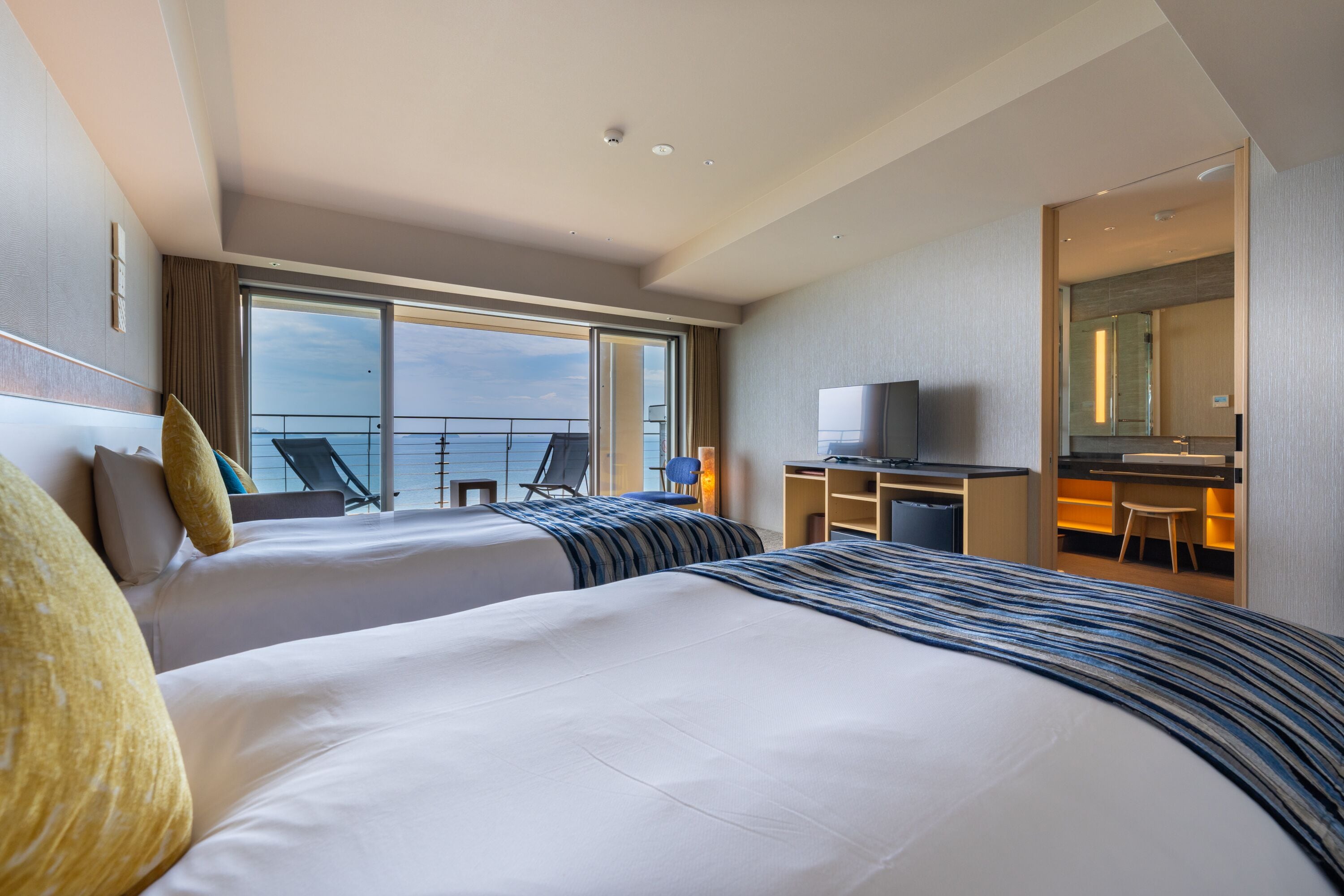 A hotel room with two beds, a desk, a television, and a view of the sea.