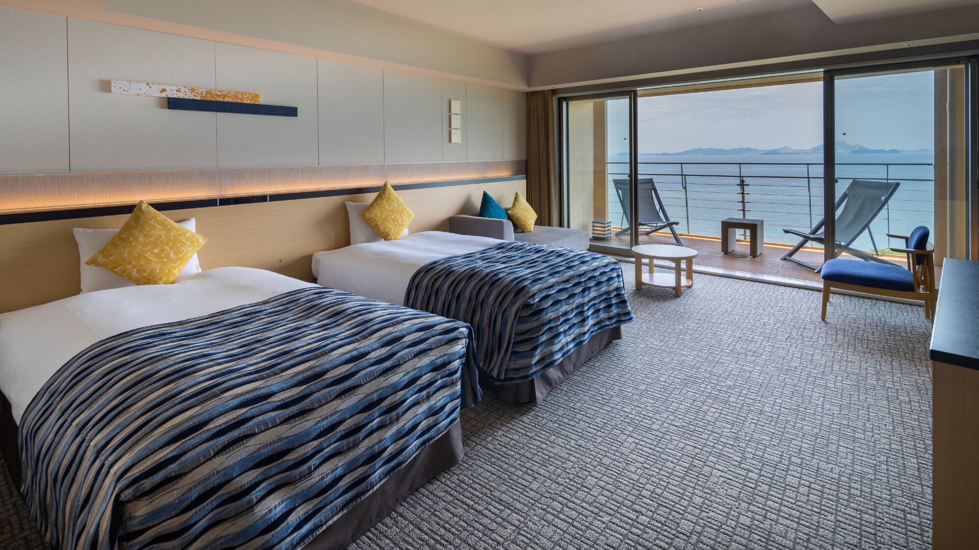 Habitación de hotel con dos camas, un balcón con vistas al mar y una pequeña mesa con sillas.
