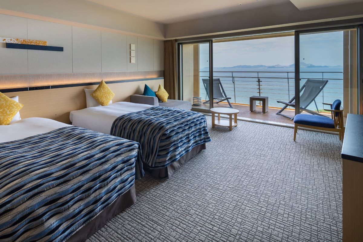 Une chambre d’hôtel avec deux lits, un balcon donnant sur la mer et une petite table avec des chaises.
