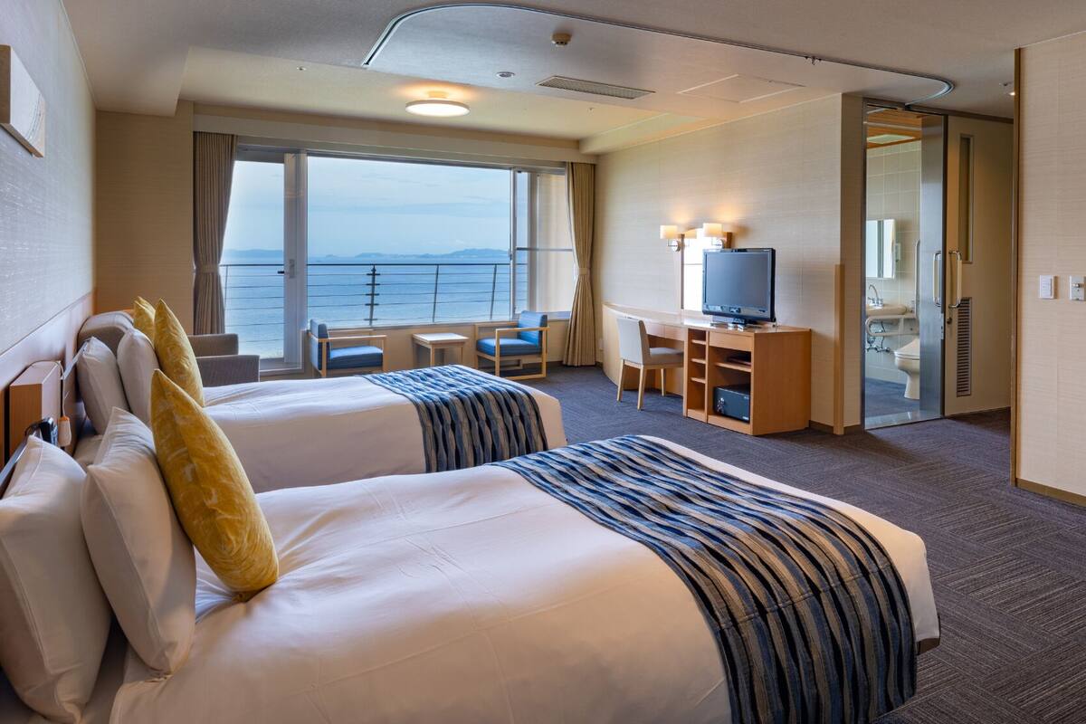 Ein Hotelzimmer mit zwei Betten, einem Schreibtisch, einem Fernseher und Meerblick.