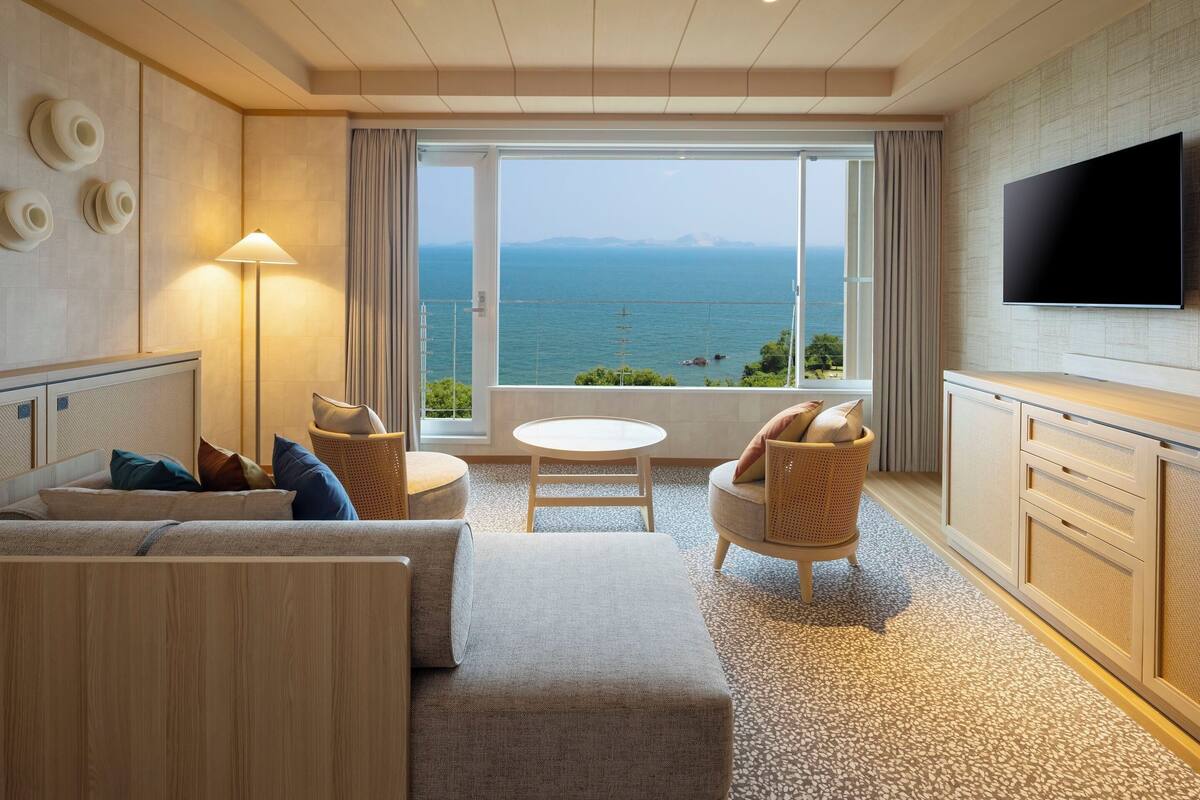 Ein Hotelzimmer mit großem Fenster und Meerblick, einem wandmontierten Flachbildfernseher, einer Couch, einem kleinen runden Tisch und einem Holzschrank.