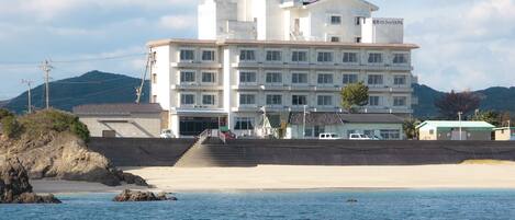 A multi-story hotel with a beach view, featuring a staircase leading to the entrance, a parking area, and a sign indicating it is a hotel.