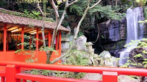 A traditional Japanese building with a tiled roof and red pillars, surrounded by lush greenery and a waterfall. - Sanage Onsen Hotel Kinsenkaku (Toyota)