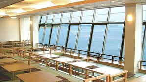 A spacious room with multiple wooden tables and benches arranged in rows, large windows offering a view of the sea. - Ikeuo-no Mifune (Minamichita)