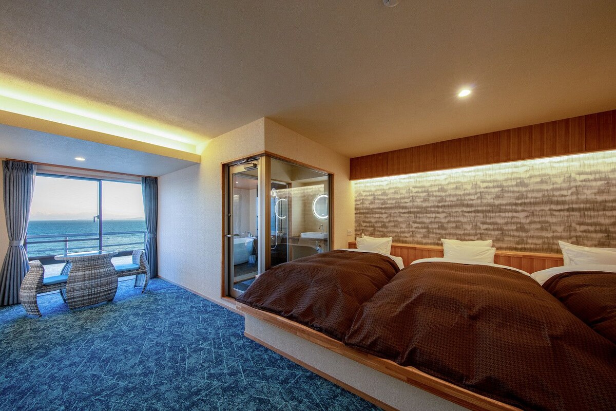 A hotel room with a large bed, a seating area with a view of the sea, a bathroom with a glass door, and a patterned headboard.