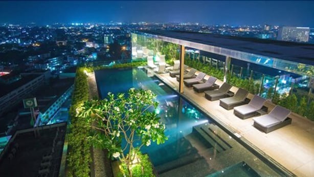 An infinity pool with lounge chairs, surrounded by greenery and overlooking a cityscape at night.