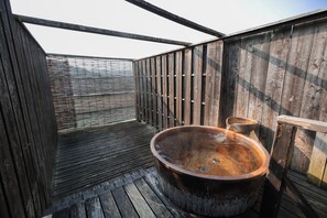 A wooden outdoor hot tub with a wooden deck and a metal fence in the background. - Yunogokan Will Be (Mimasaka)
