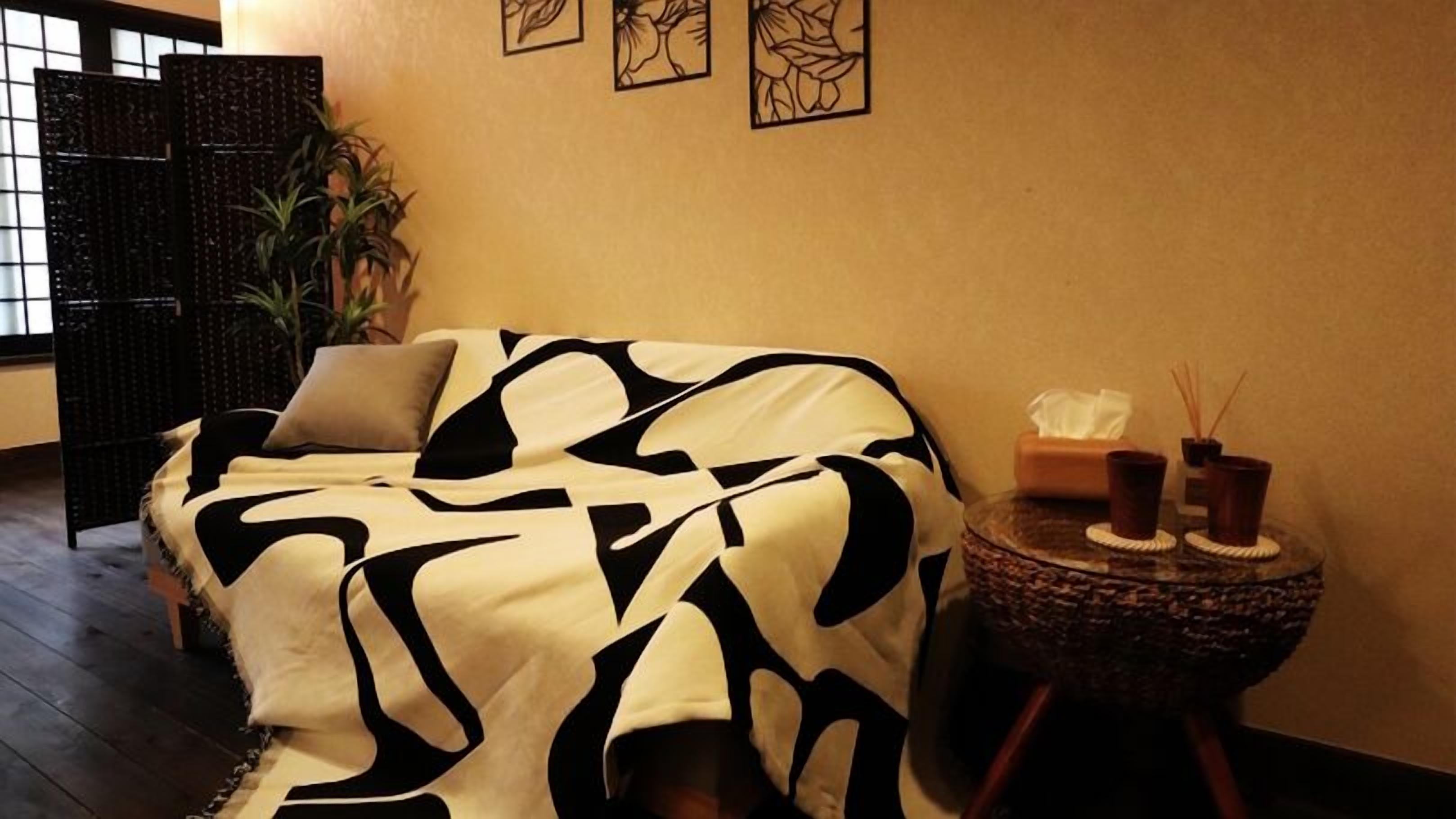 A room with a sofa covered in a black and white patterned blanket, a small round side table with candles and a tissue box, and a potted plant.