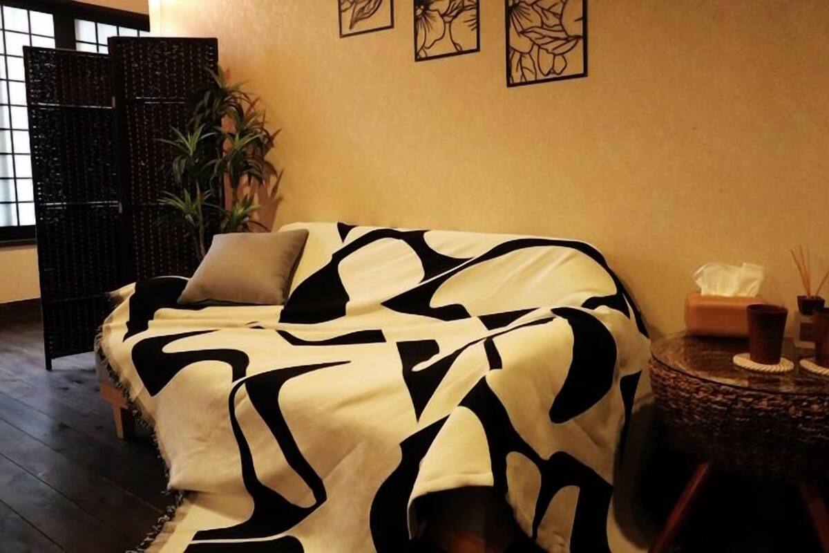 A room with a sofa covered in a black and white patterned blanket, a small round side table with candles and a tissue box, and a potted plant.