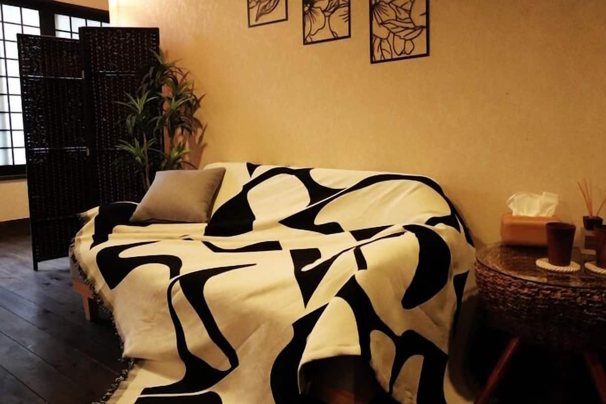 A room with a sofa covered in a black and white patterned blanket, a small round side table with candles and a tissue box, and a potted plant.