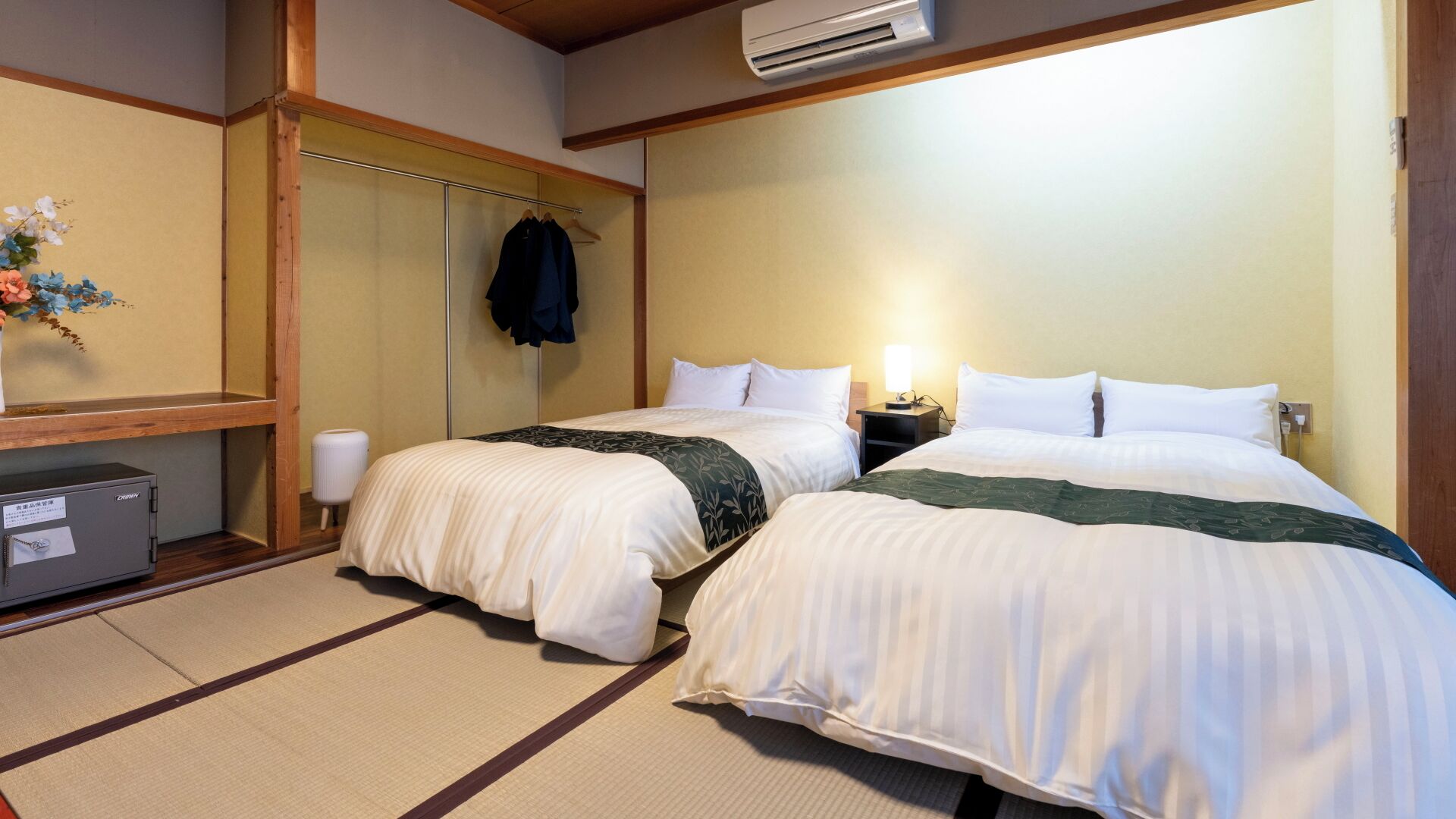 A hotel room with two beds, a wardrobe, a bedside table with a lamp, and an air conditioning unit.