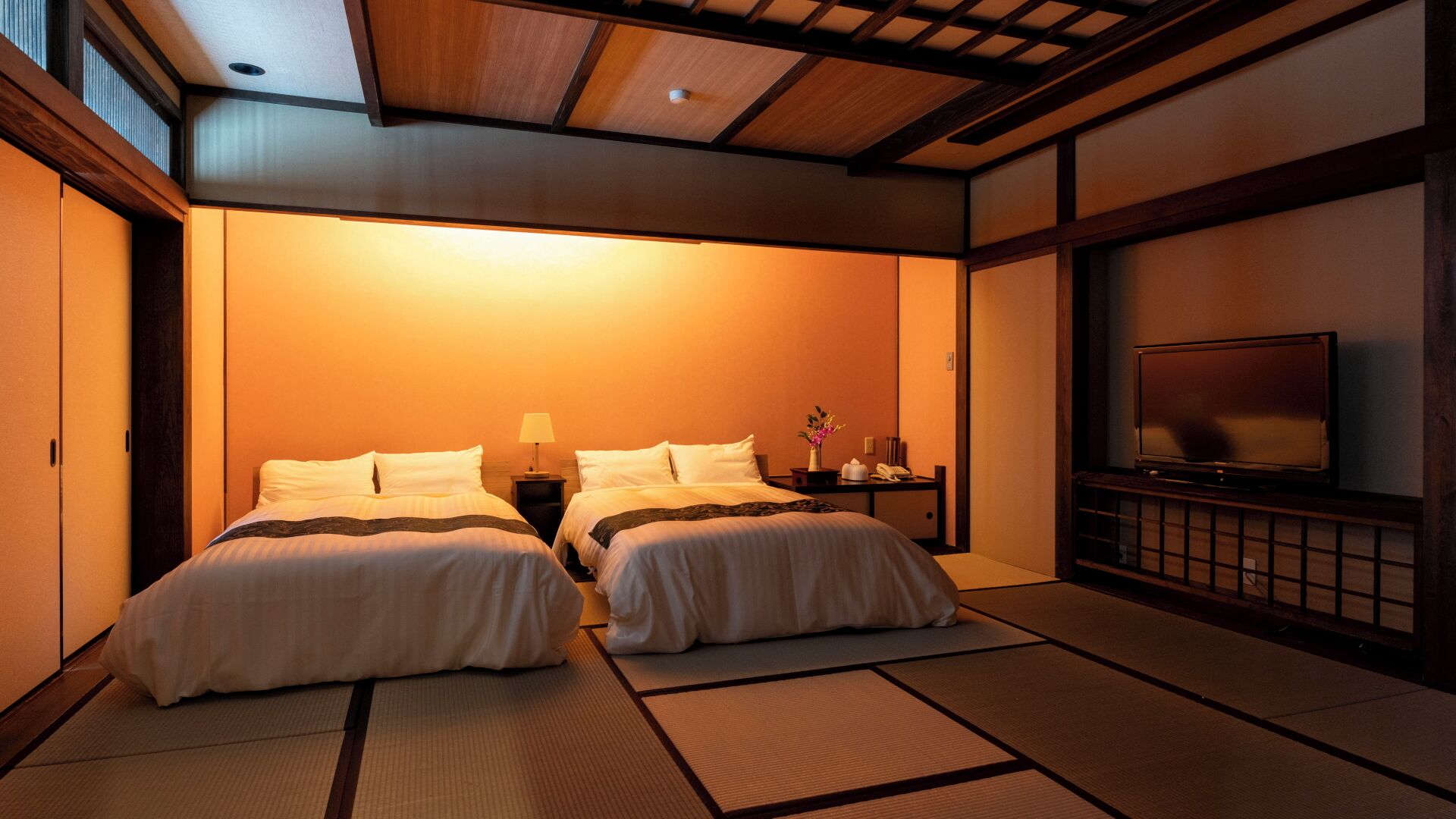 A traditional Japanese-style room with tatami flooring, two beds, a television, and a small table with a lamp and phone.