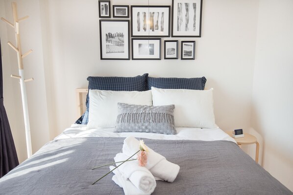 A neatly made bed with a grey and white checkered duvet, a grey pillow, and a white pillow. There are towels and a flower on the bed. A wooden coat rack is to the left of the bed.