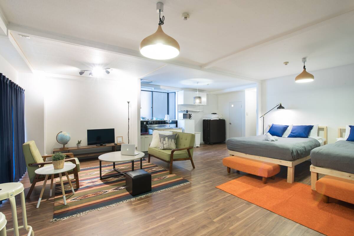 Una sala de estar moderna con un sofá, una mesa de centro y un televisor. Se observa una cocina al fondo con armarios y un refrigerador.