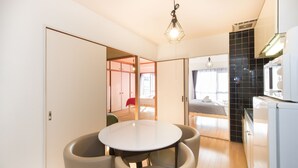 A modern kitchen with a round dining table and chairs, a hanging light fixture, and a view into a bedroom through an open door. - Namba Hara Apartment (Osaka)