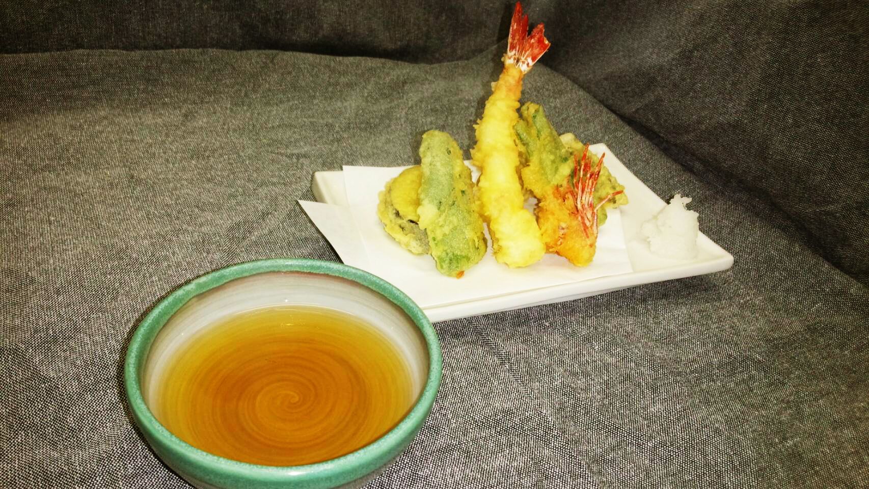 A plate of tempura shrimp and vegetables on a gray couch with a bowl of dipping sauce.