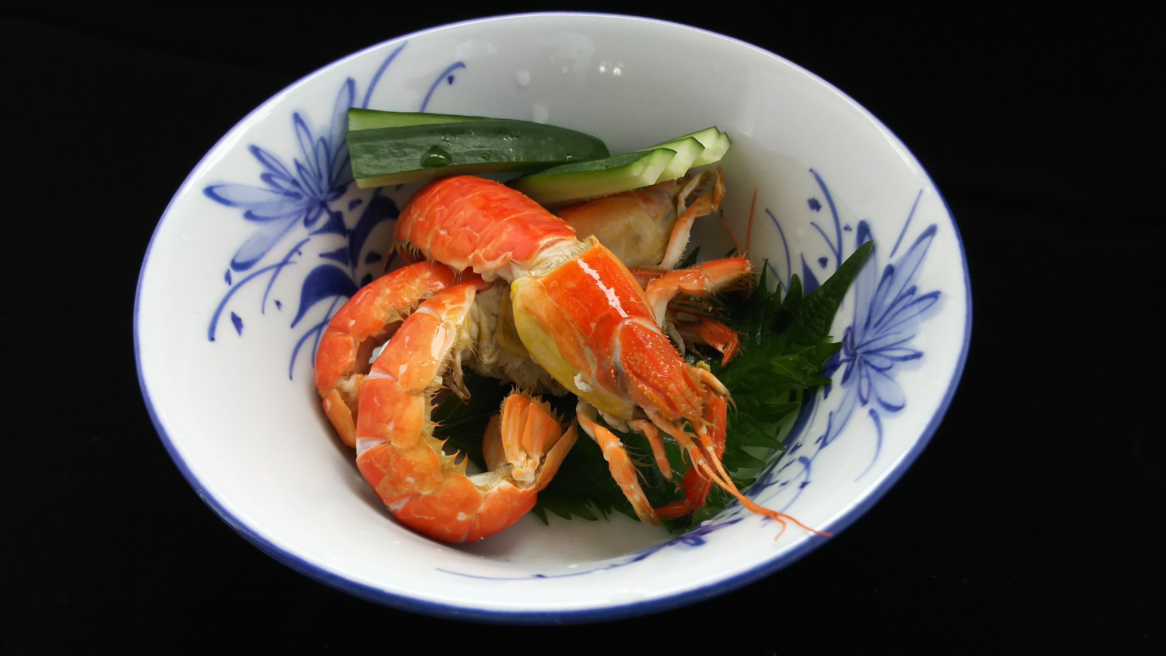 A bowl with a floral pattern containing a crab, cucumber slices, and green leaves.