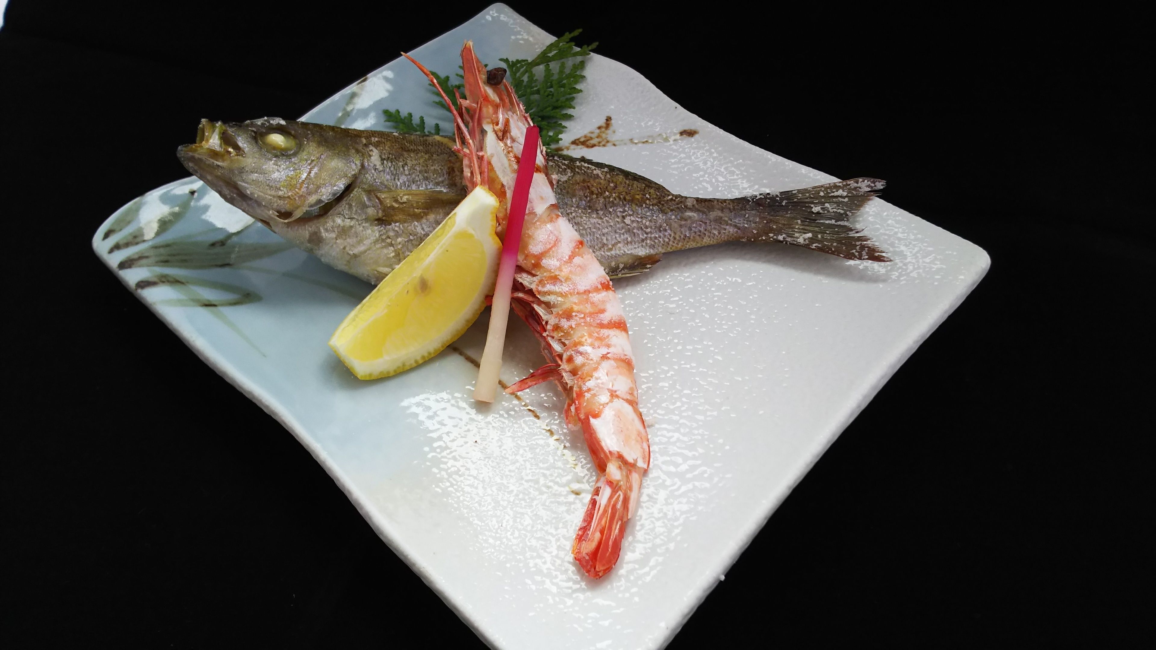A plate with a whole fish, a large shrimp, and a lemon wedge.