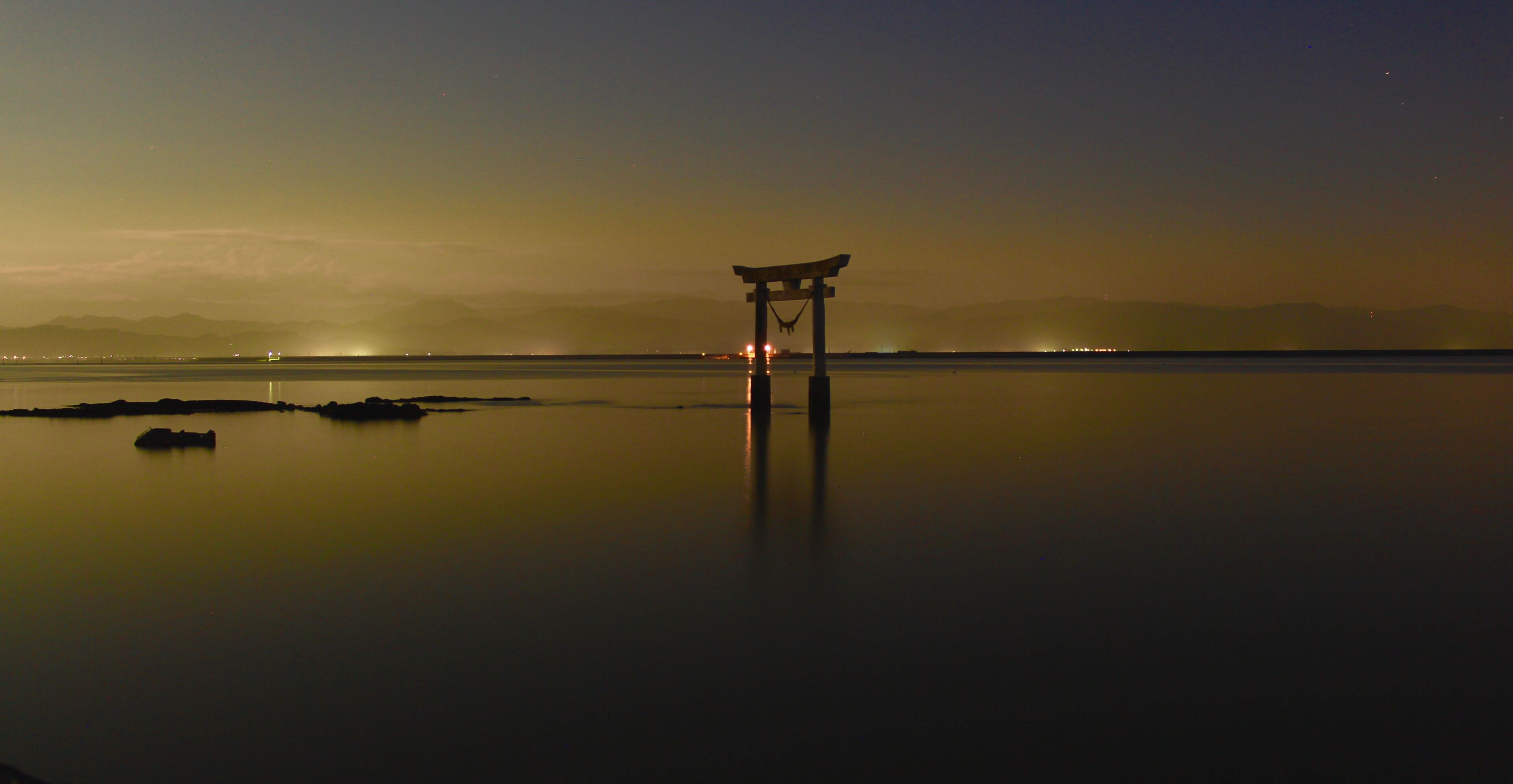 A serene nighttime scene with a traditional Japanese torii gate in the foreground, reflecting on a calm body of water.