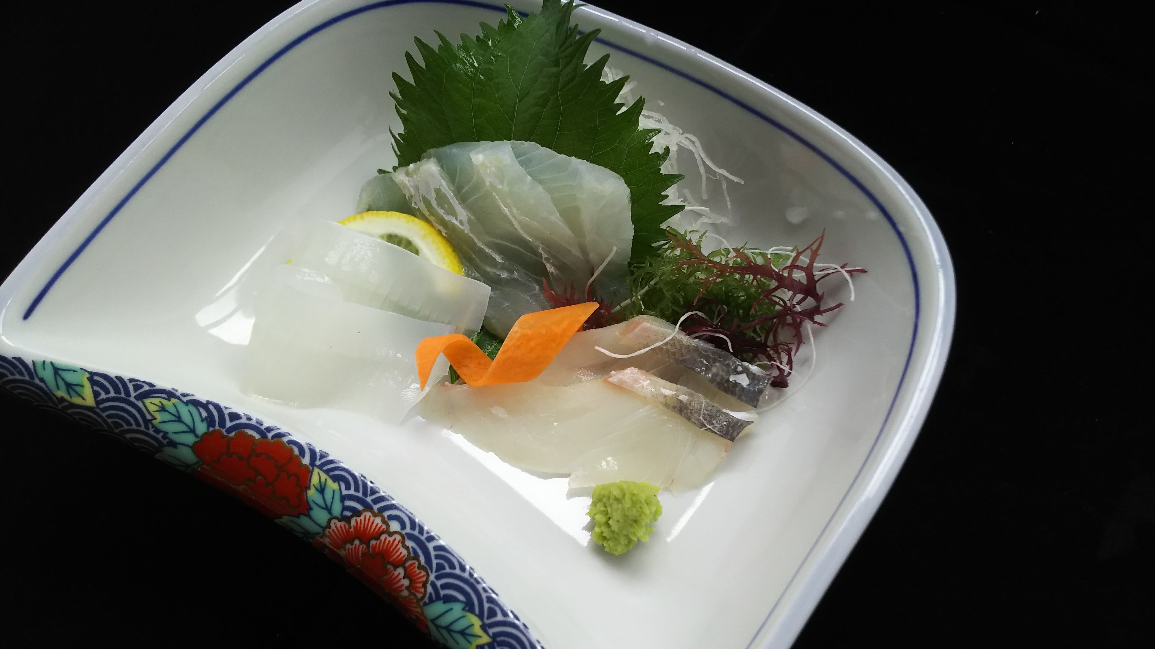 En firkantet, hvid tallerken med blå kant, der indeholder sashimi, en skive citron, en gulerodsstykke og grønt.