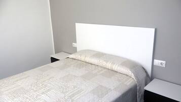 Una cama con cabecera blanca en una habitación con pared lisa y una mesita de noche.