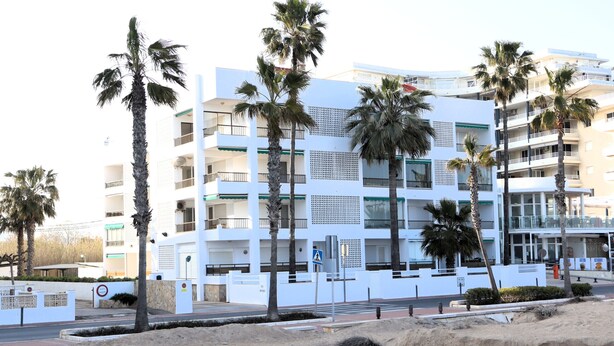A multi-story building with balconies, palm trees, and a sandy foreground. - Los Monegros (Peñiscola)