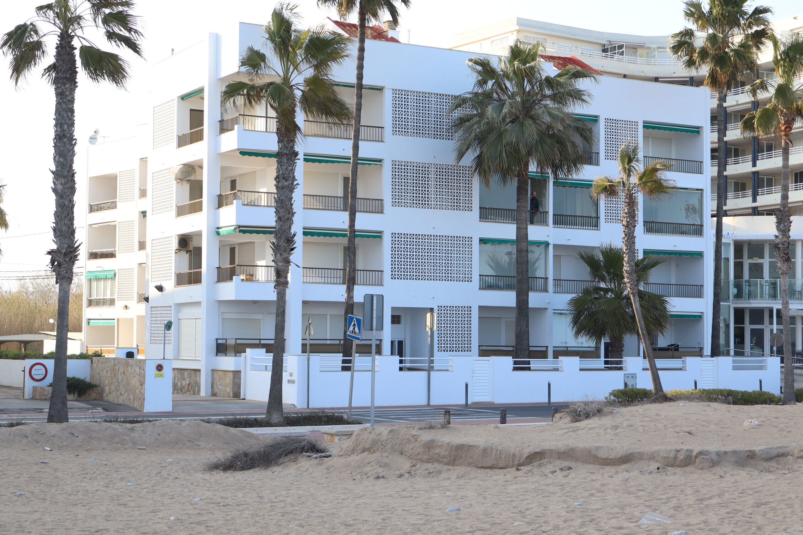 A multi-story building with balconies, palm trees, and a sandy foreground.