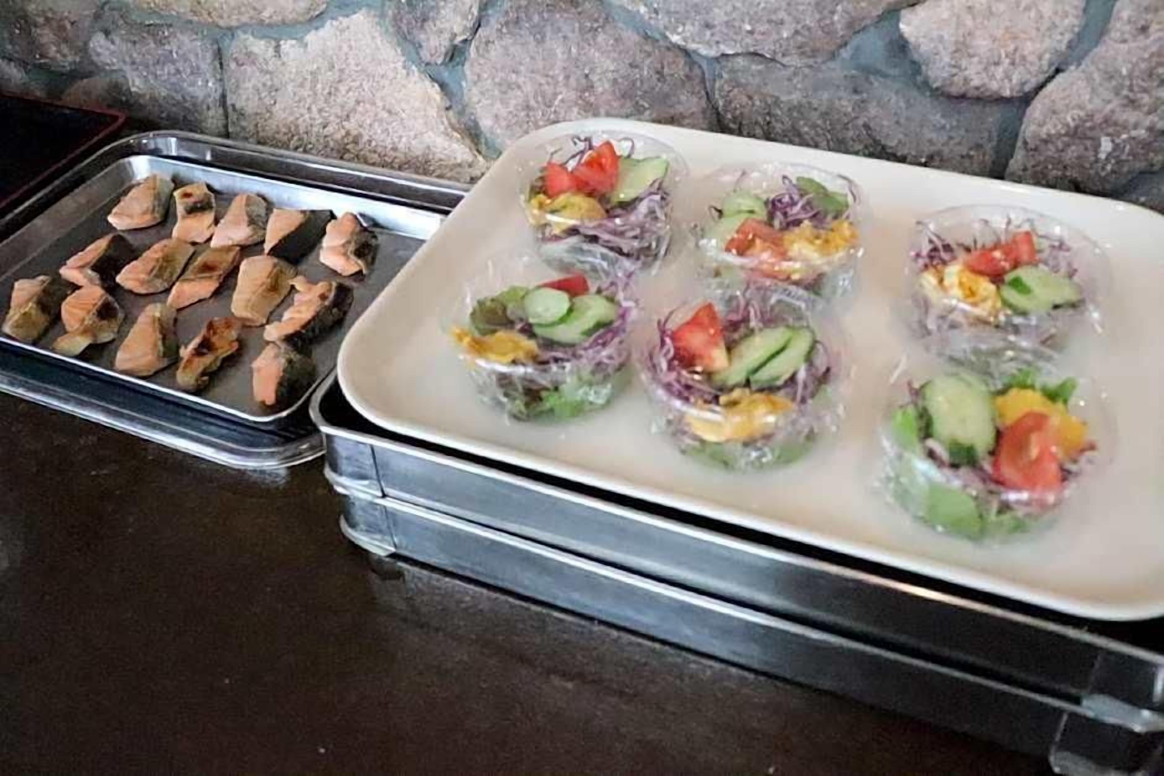 A tray of food items, including what appears to be grilled fish and a variety of vegetable and fruit salads.