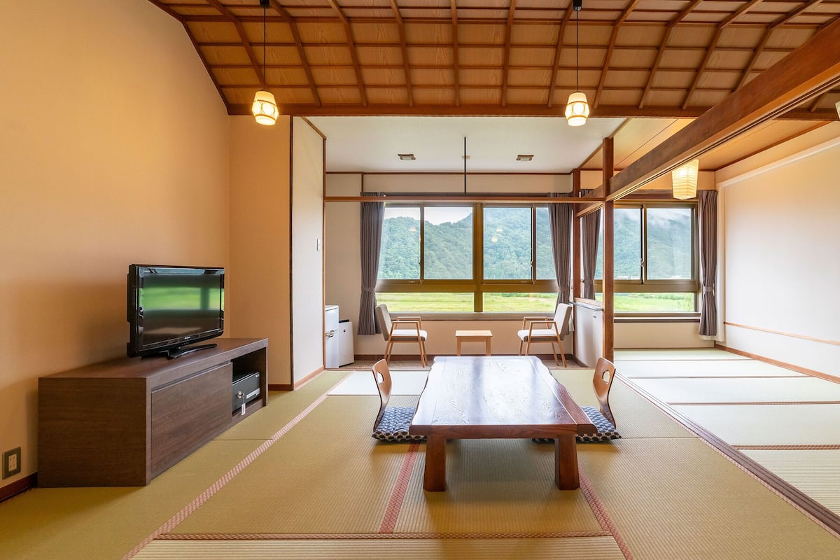 A traditional Japanese room with tatami flooring, a television, a wooden table, and a view of a mountain landscape through large windows.