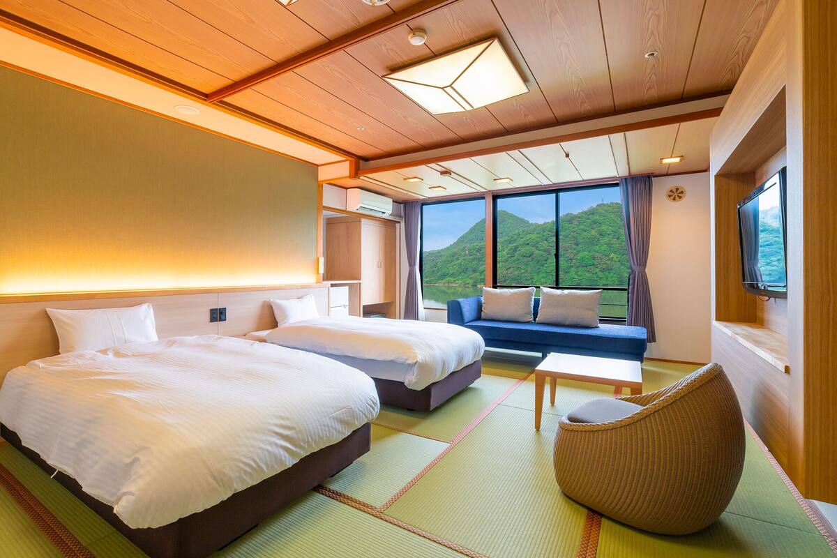 A hotel room with two beds, a sofa, a TV, and a view of a green landscape.