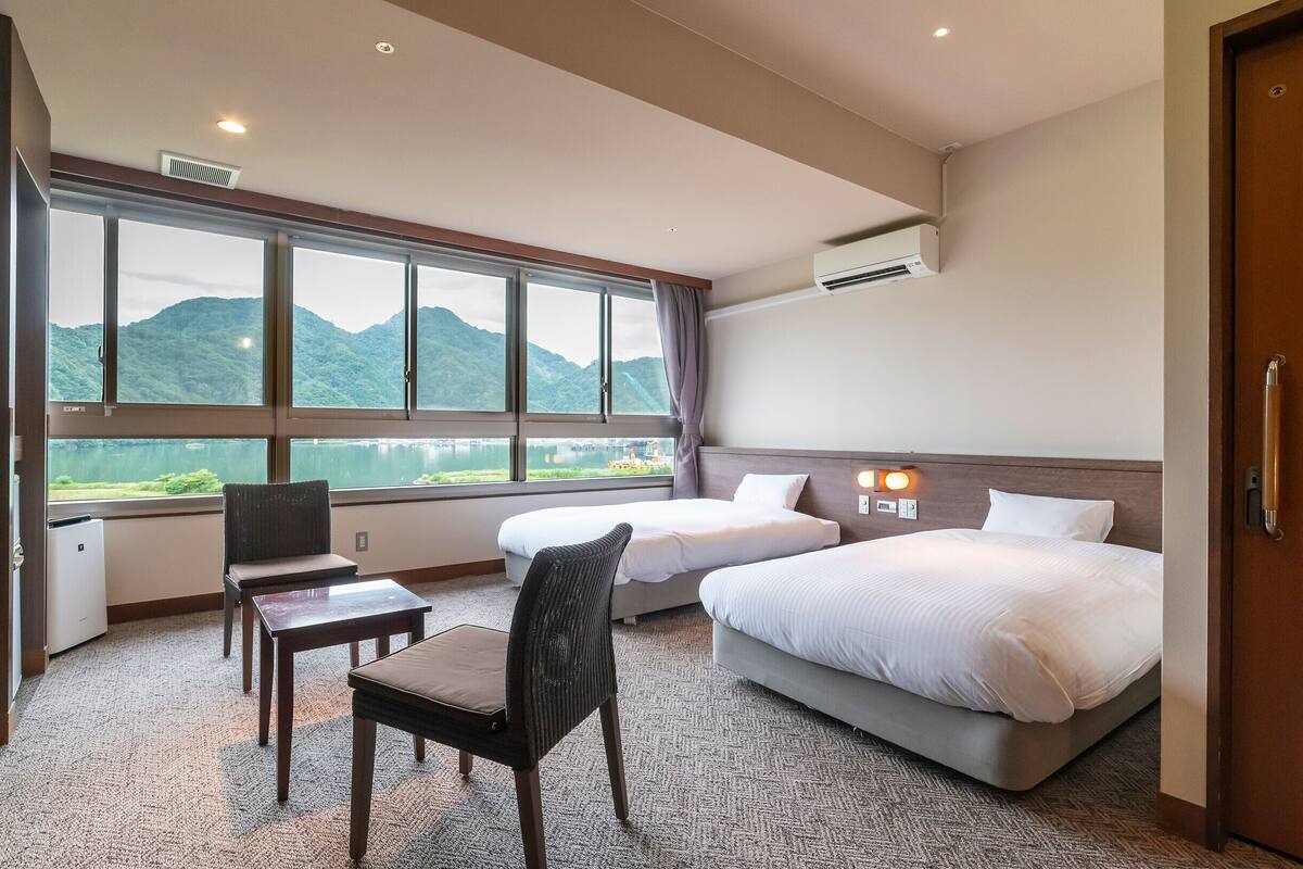 A hotel room with two beds, a desk, a chair, and a large window offering a view of mountains and a lake.