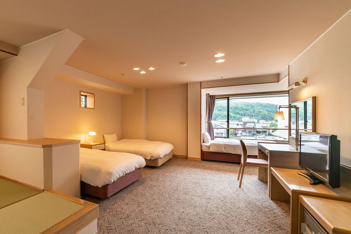 A hotel room with two beds, a desk, a chair, a TV, and a window with a view of buildings.