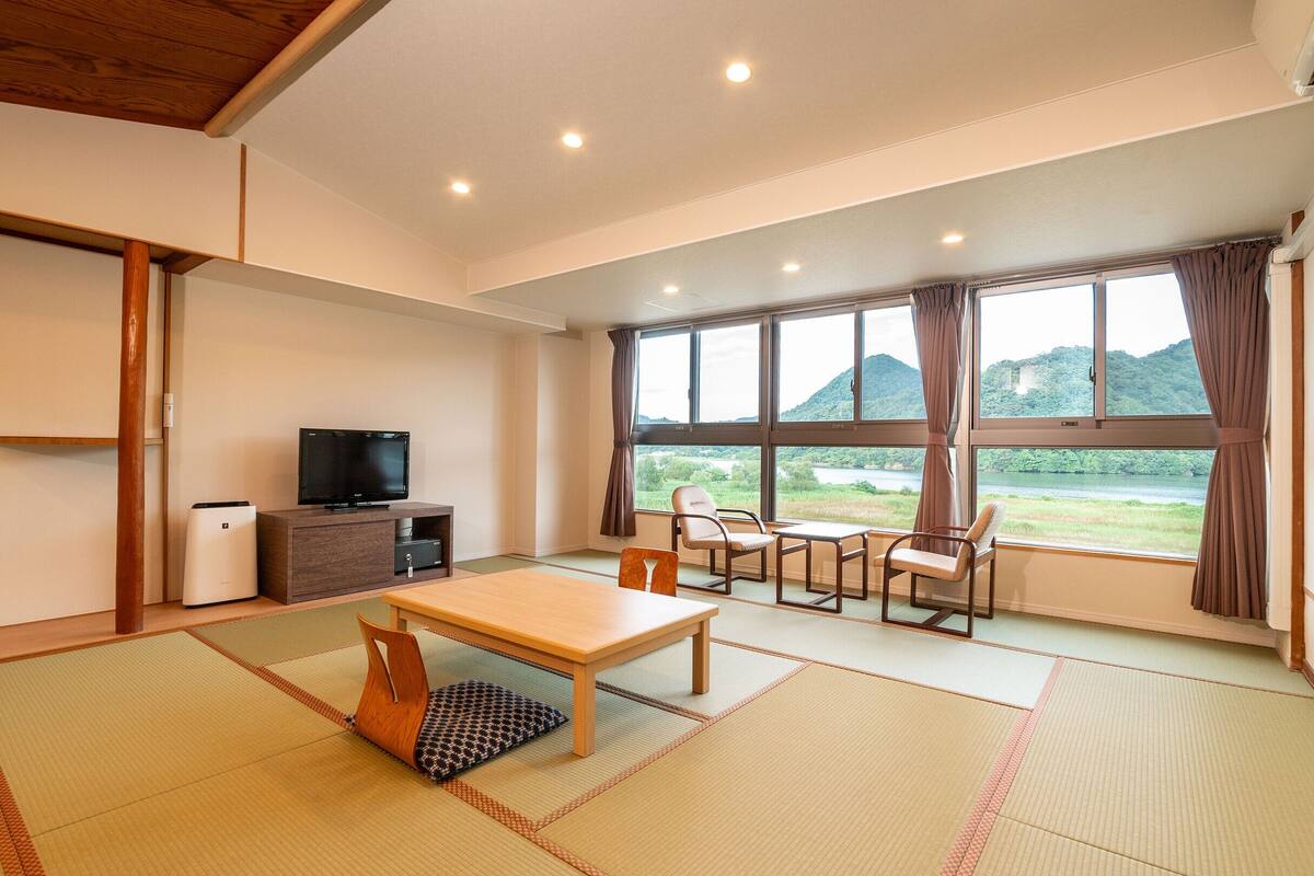 A room with tatami flooring, a television, a table, chairs, and a view of a lake and mountains through large windows.