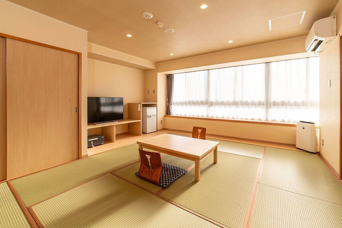 A traditional Japanese-style room with tatami flooring, a low table, a television, a small refrigerator, and a window with curtains.