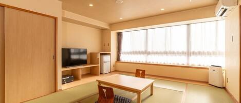 A traditional Japanese-style room with tatami flooring, a low table, a television, a small refrigerator, and a window with curtains.