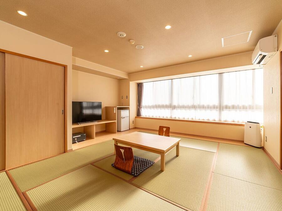 Une pièce de style japonais traditionnel, avec un sol recouvert de tatamis, une petite table basse, un téléviseur, un petit réfrigérateur et une fenêtre avec des rideaux.