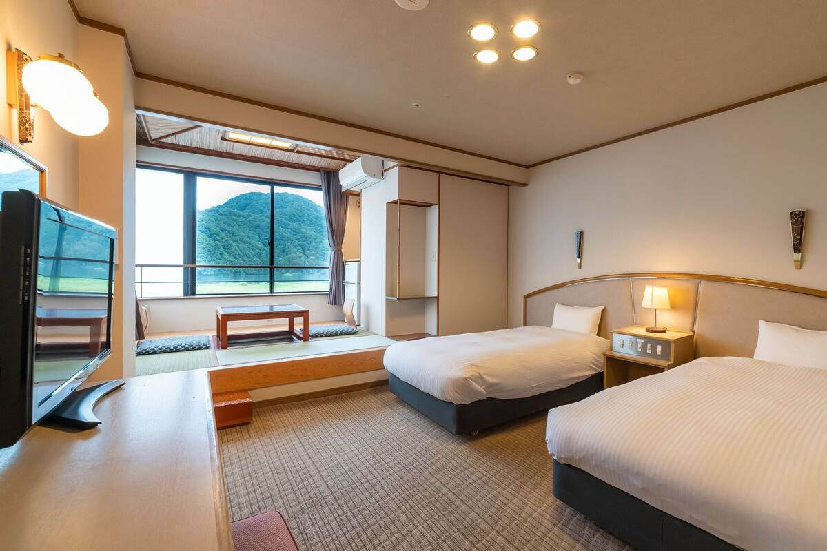 A hotel room with two beds, a desk, a television, and a window with a view of a mountain.