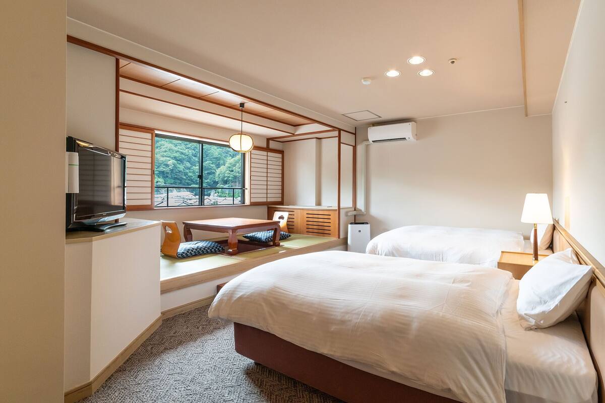 A hotel room with two beds, a desk, a TV, and a window with a view of trees.