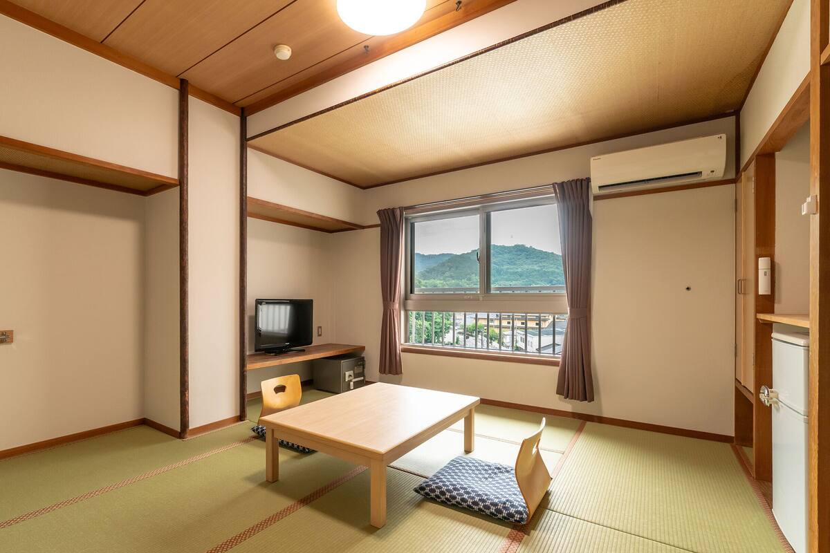 A room with tatami flooring, a wooden table, a television, and a window with a view of hills.