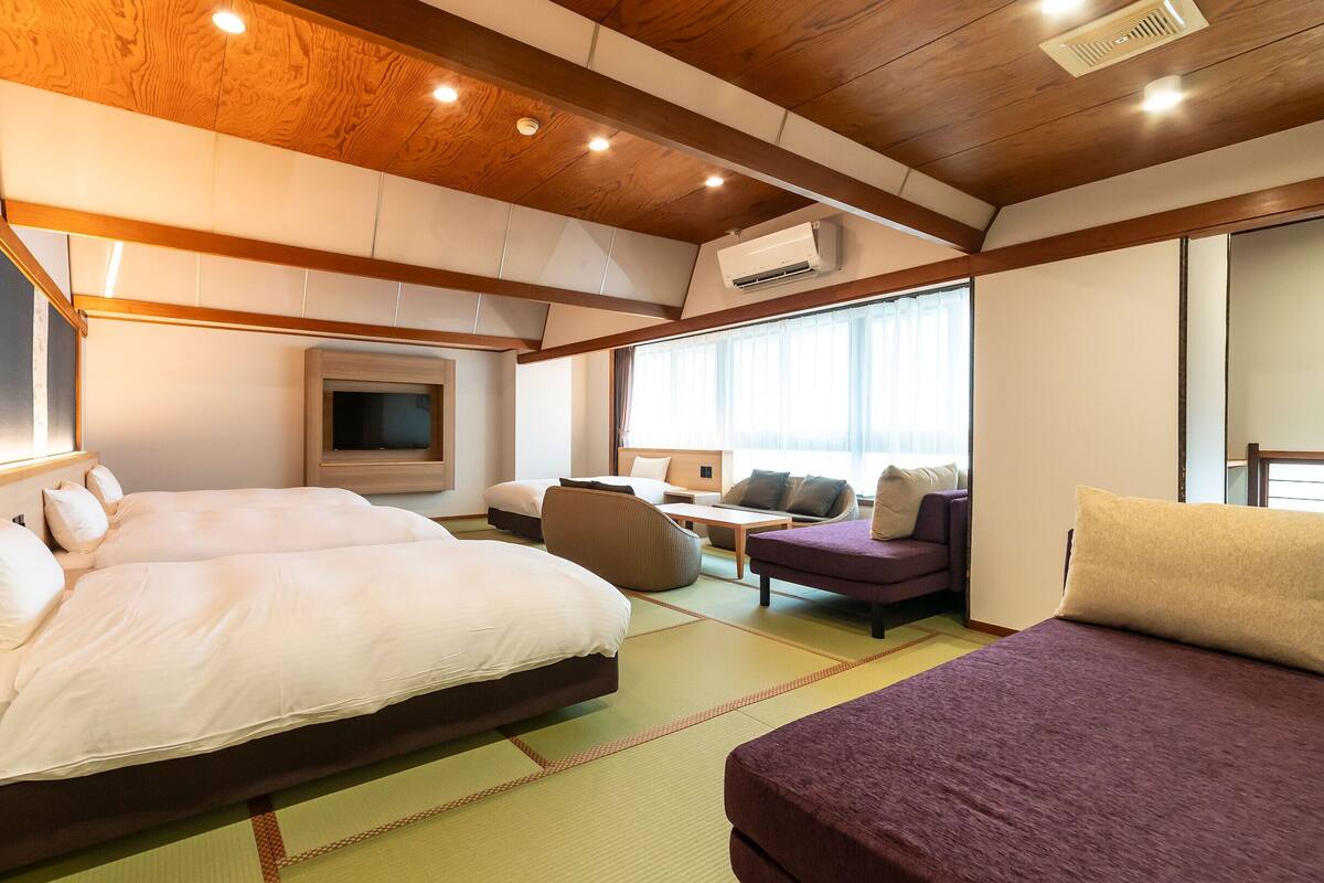 A hotel room with a wooden ceiling, a bed, a sofa, a TV, and a large window.