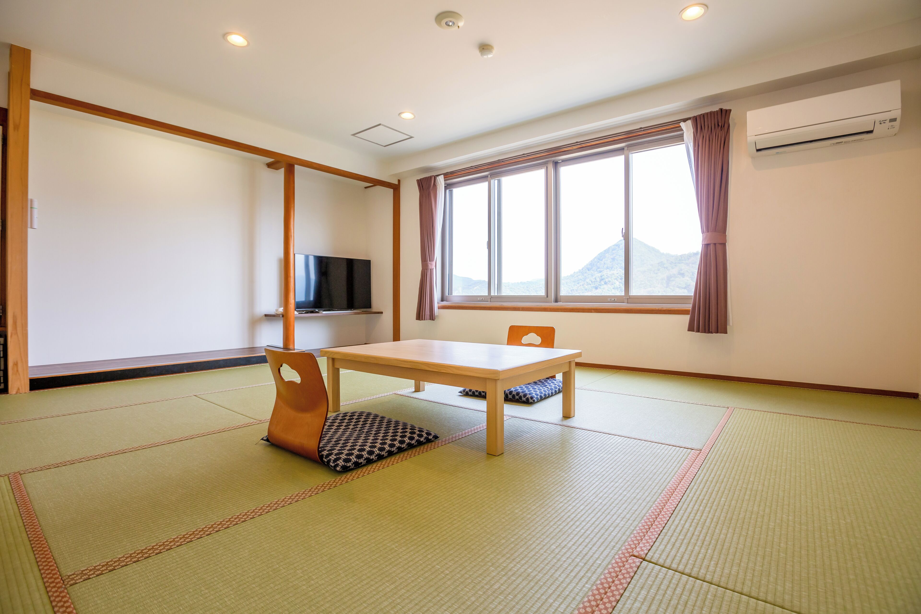 A room with tatami flooring, a wooden table, a television, and a window with a view of a mountain.