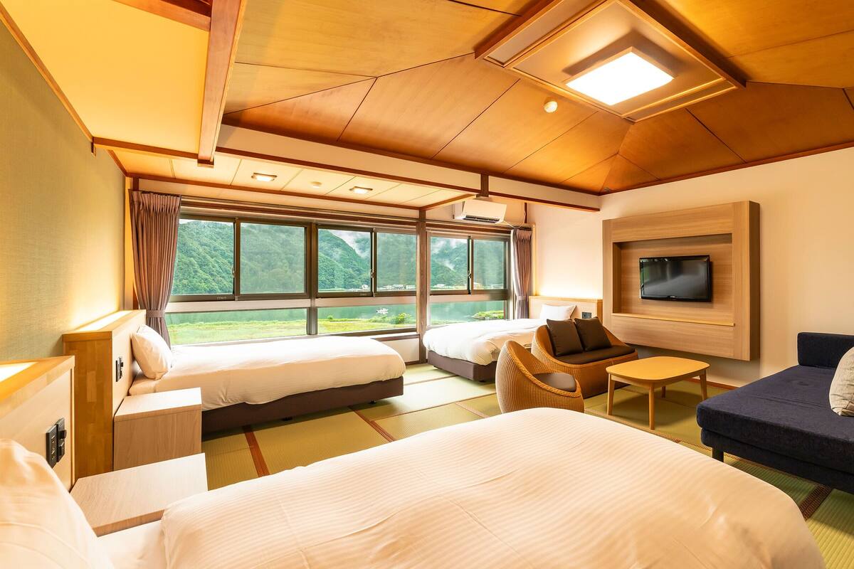 A hotel room with a large bed, a sofa, a TV mounted on the wall, and a window offering a view of greenery.